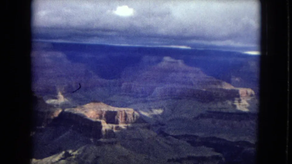 1967 home-movie frame looking across the Grand Canyon toward a distant rim under heavy cloud cover, with soft grain and a blue-purple color cast.