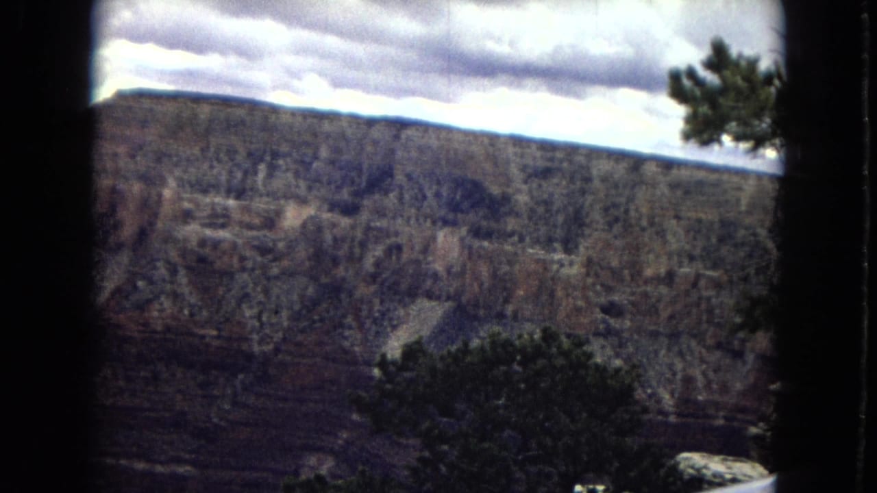 1967 8mm view of the Grand Canyon in Arizona, showing a broad cliff face under heavy clouds with dark foreground trees near the rim.