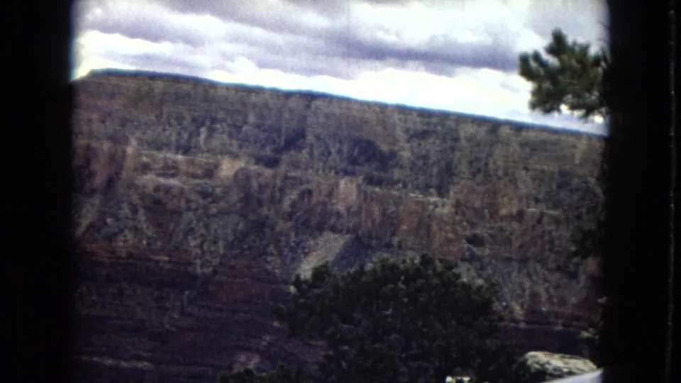 1967 8mm view of the Grand Canyon in Arizona, showing a broad cliff face under heavy clouds with dark foreground trees near the rim.