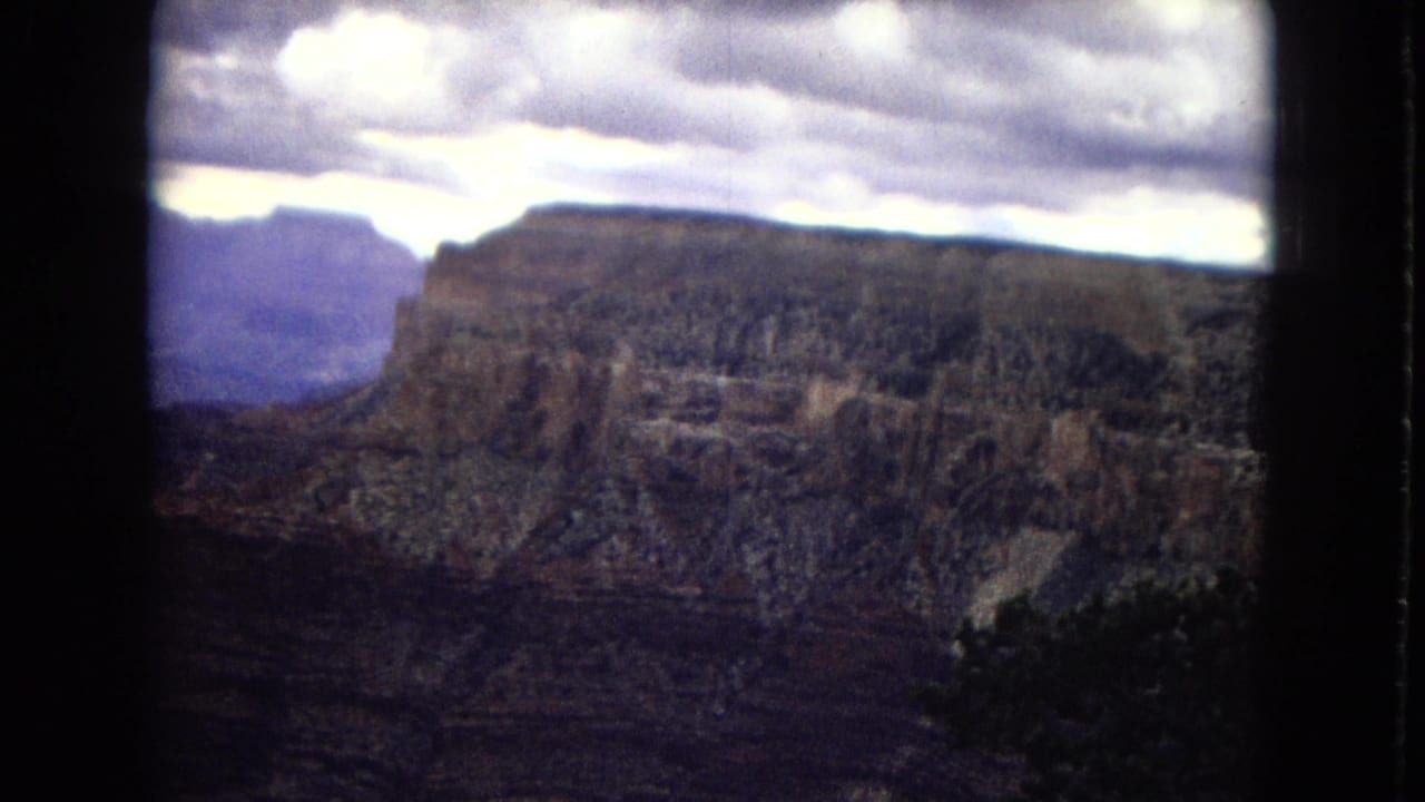 1967 8mm Grand Canyon panorama with a flat-topped cliff, distant rock formations, and a bright cloudy sky above the canyon.