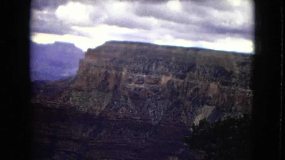 1967 8mm Grand Canyon panorama with a flat-topped cliff, distant rock formations, and a bright cloudy sky above the canyon.