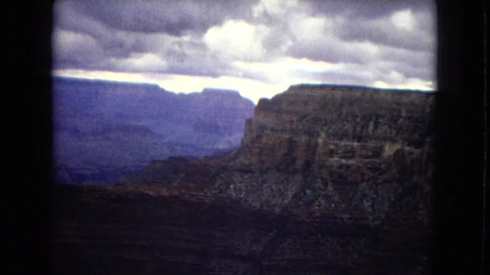 1967 Arizona home-movie frame of the Grand Canyon, with a dark foreground rim, distant blue-toned cliffs, and heavy cloud cover.