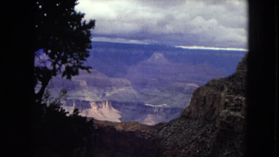 1967 8mm view across the Grand Canyon in Arizona, with a dark tree silhouette at the rim and distant canyon layers under a cloudy sky.