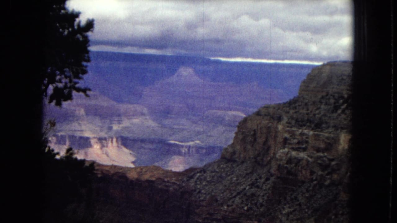 1967 Arizona 8mm frame showing Grand Canyon rock formations and distant mesas through a dark window-like frame, with heavy clouds above.