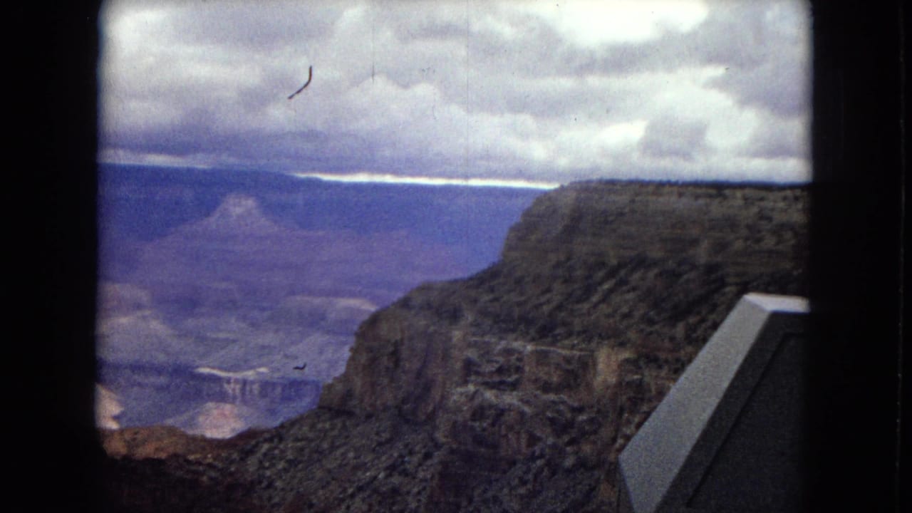 1967 Arizona 8mm frame of the Grand Canyon under overcast sky, framed by dark vertical edges with a partial angular overlook element at the right side.