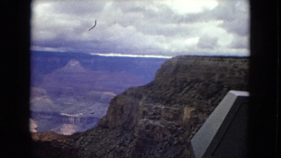 1967 Arizona 8mm frame of the Grand Canyon under overcast sky, framed by dark vertical edges with a partial angular overlook element at the right side.
