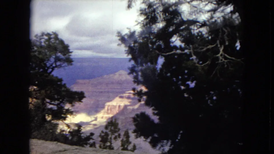 1967 Arizona 8mm frame showing a Grand Canyon vista partly framed by dark tree silhouettes, with pale rock layers and a cloudy sky beyond.