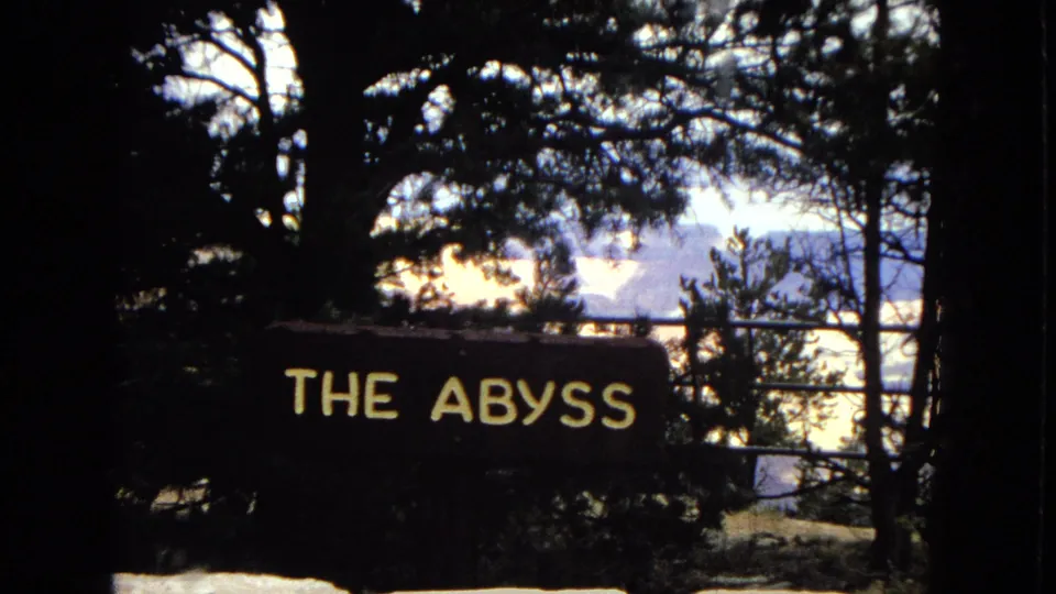 1967 Arizona 8mm frame showing a dark overlook scene with a readable "THE ABYSS" sign, tree silhouettes, and the Grand Canyon visible in the distance.