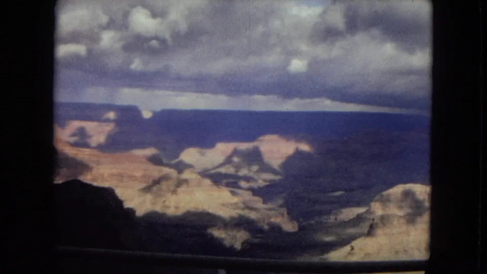 1967 Arizona 8mm frame showing a wide Grand Canyon view with layered rock, stormy clouds, and an overlook railing along the lower edge.