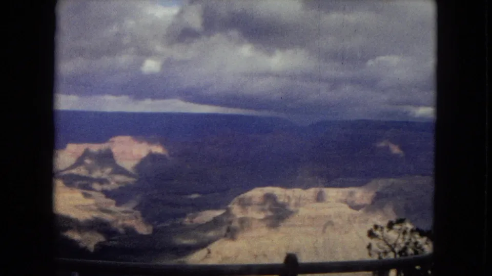 1967 Arizona 8mm frame of the Grand Canyon from an overlook, with broad canyon walls, a cloudy sky, a railing, and small branches in the lower right.