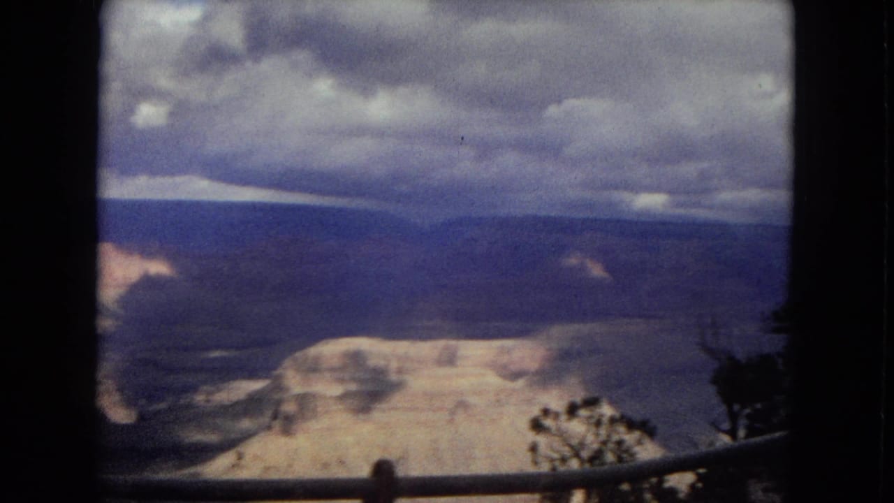 1967 8mm Grand Canyon overlook view with a dark railing in the foreground, cloudy sky, and distant layered canyon walls.