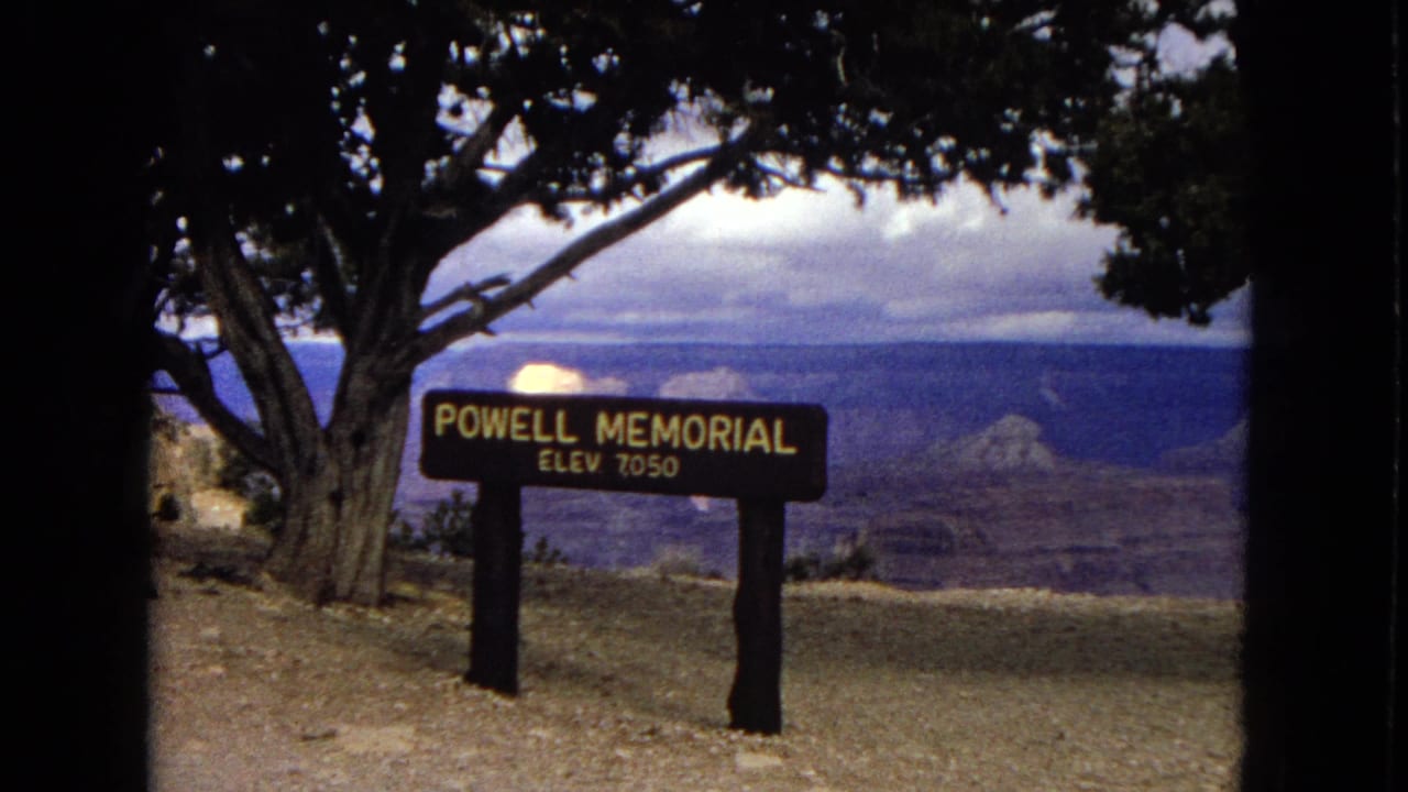 Powell Memorial sign marked elevation 7050 at a Grand Canyon overlook, with trees, dirt ground, and canyon ridges in the background.