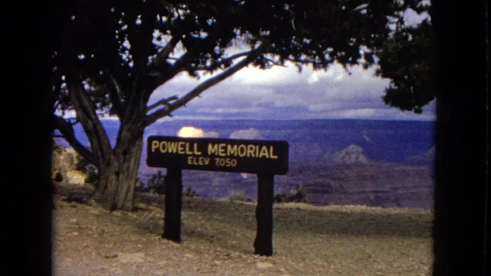 Powell Memorial sign marked elevation 7050 at a Grand Canyon overlook, with trees, dirt ground, and canyon ridges in the background.