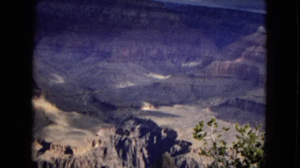 Wide 1967 Grand Canyon vista with foreground shrubs, pale rock shelves, shadowed canyon depth, and a cloudy sky.