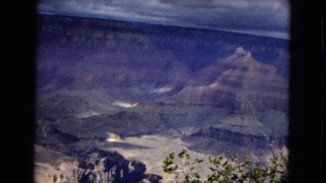 1967 8mm Arizona travel footage showing a broad Grand Canyon view, layered rock walls, distant ridges, and foreground shrubs beneath a moody sky.