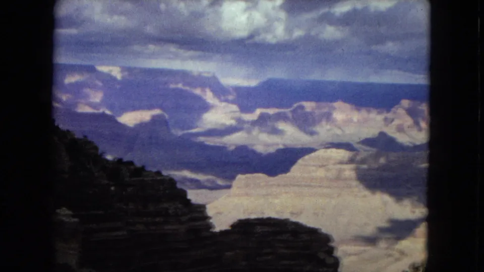 1967 8mm Arizona footage looking across the Grand Canyon from a high overlook, with a shadowed cliff in front and pale canyon formations in the distance.