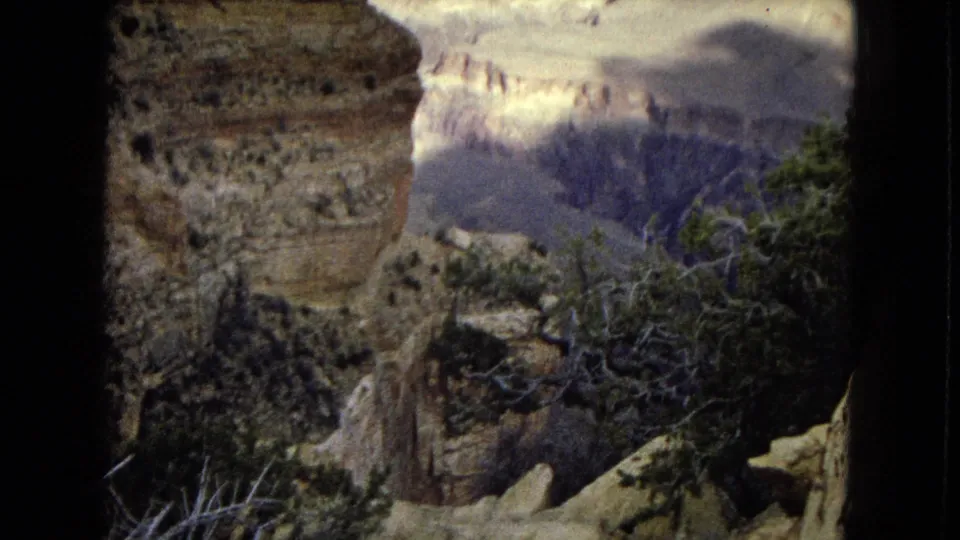 1967 8mm Arizona footage from a canyon rim, with a close rocky wall, desert shrubs, and distant Grand Canyon cliffs partly softened by haze and exposure shift.