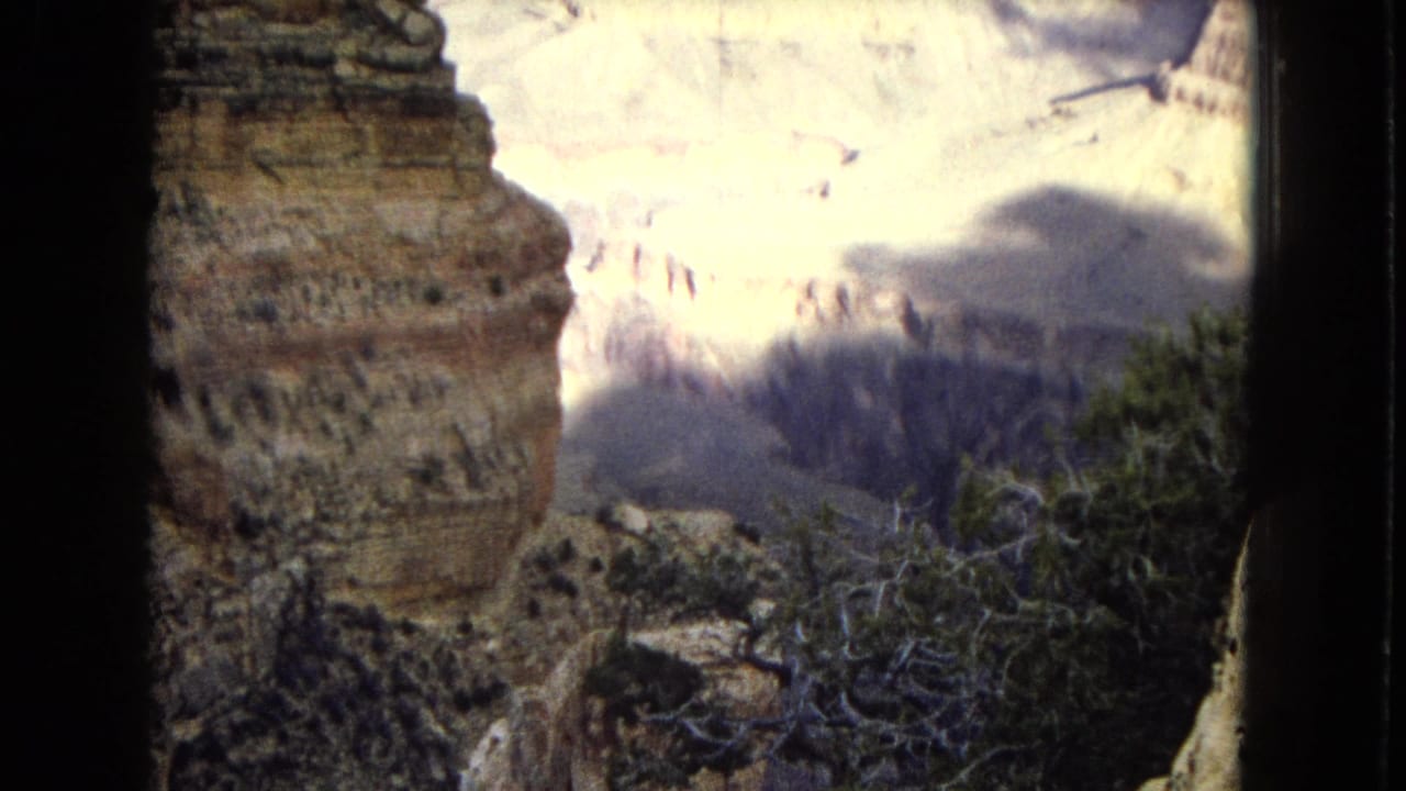1967 8mm home-movie frame of the Grand Canyon area showing a close rock wall, scrubby foreground vegetation, distant pale canyon layers, and a large cloud shadow.