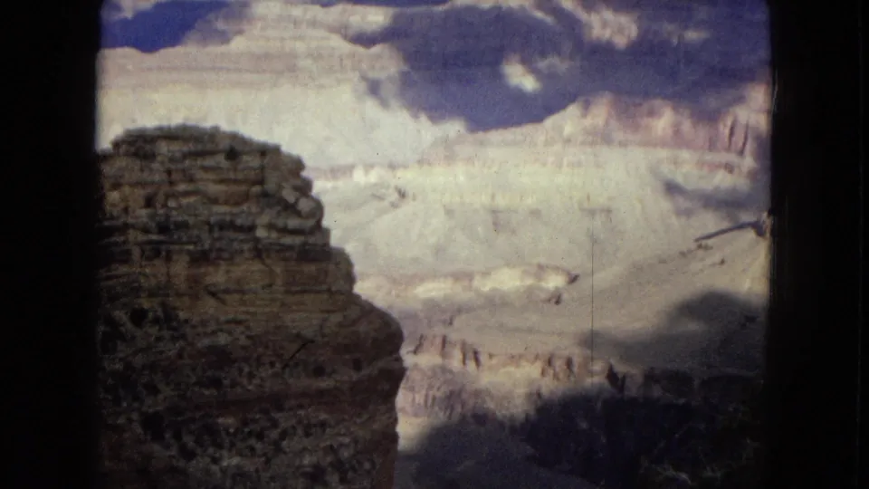 1967 8mm home-movie frame looking across the Grand Canyon, with a dark foreground rock formation, pale stratified canyon walls, blue sky, and dramatic clouds.
