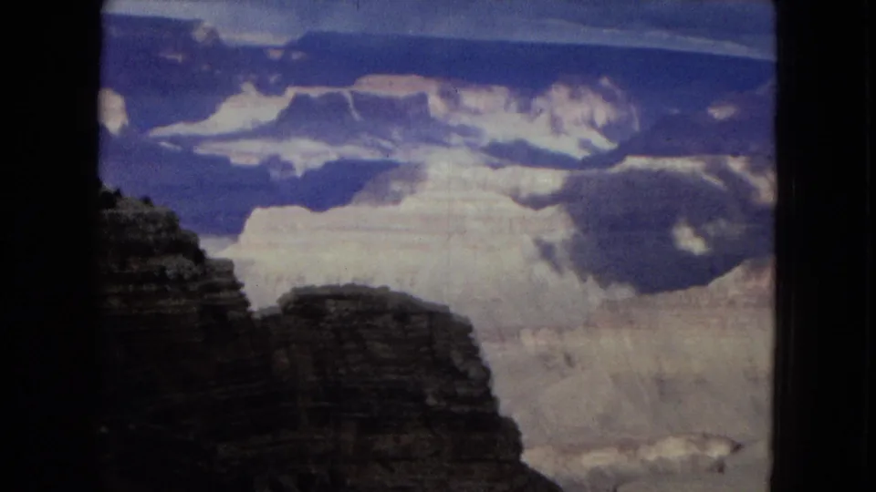 1967 8mm home-movie frame showing a shadowed canyon rim in the foreground, bright layered canyon walls in the distance, and heavy clouds across a blue sky.