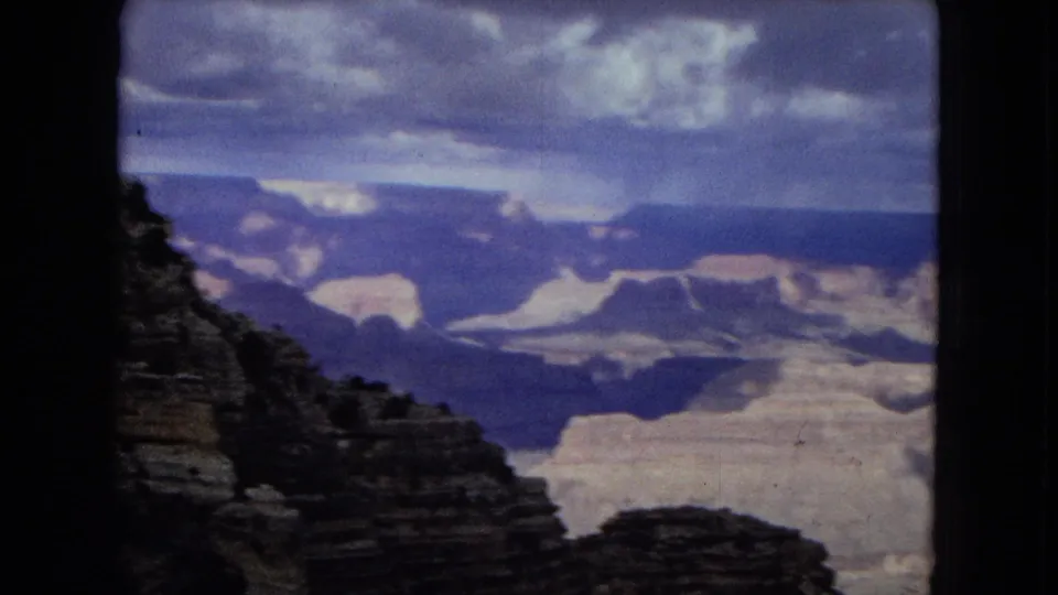 1967 8mm home-movie frame of a wide Grand Canyon view, with dark foreground cliffs, sunlit distant formations, blue sky, and broken clouds over the canyon.