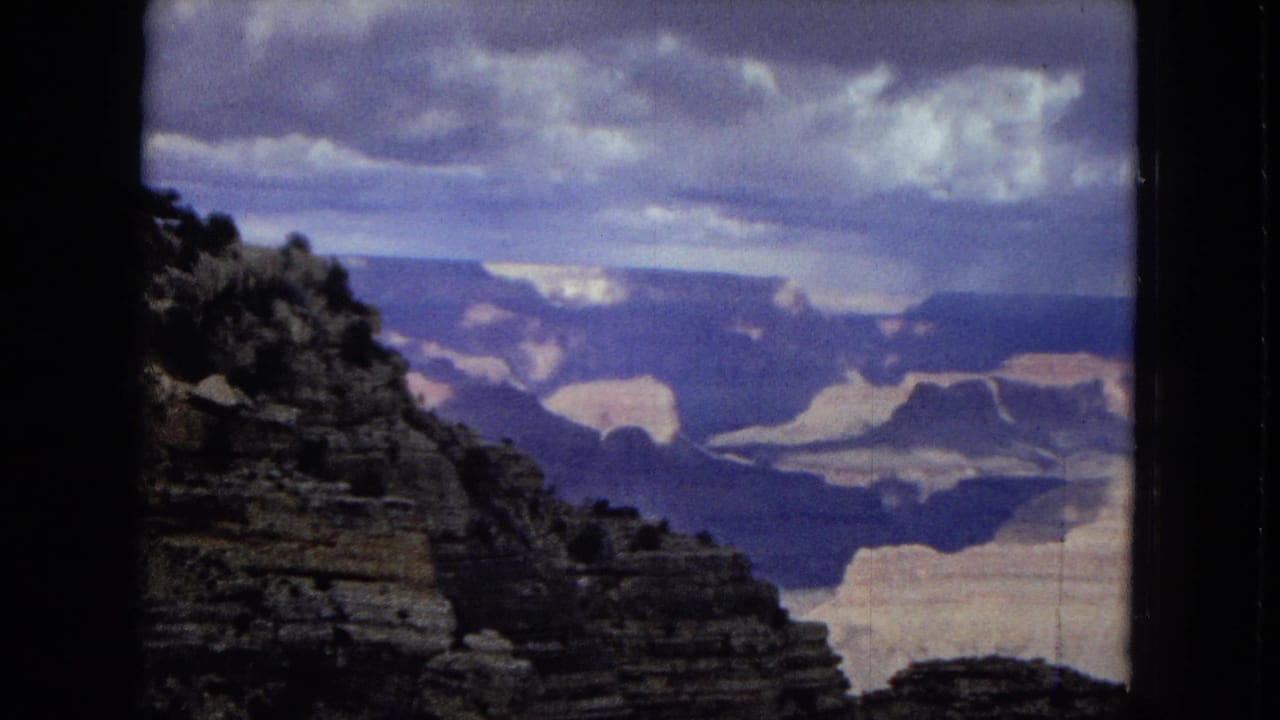 1967 8mm home-movie view across the Grand Canyon, with layered canyon walls, deep ravines, a cloudy sky, and dark foreground rock.