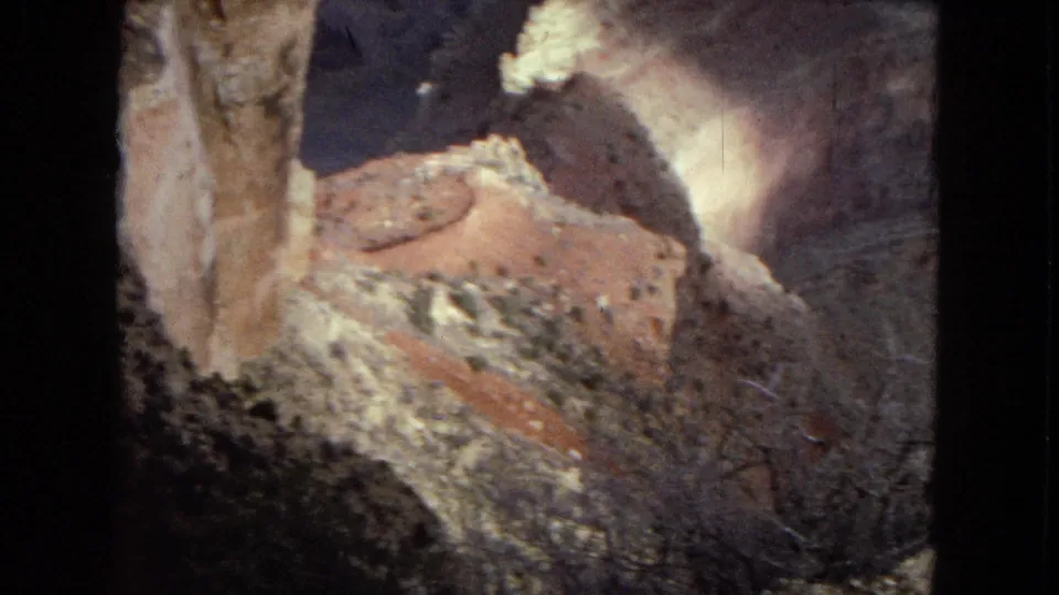 1967 8mm Grand Canyon footage showing a reddish slope, pale cliff wall, dark canyon shadow, and rough foreground terrain from an elevated viewpoint.