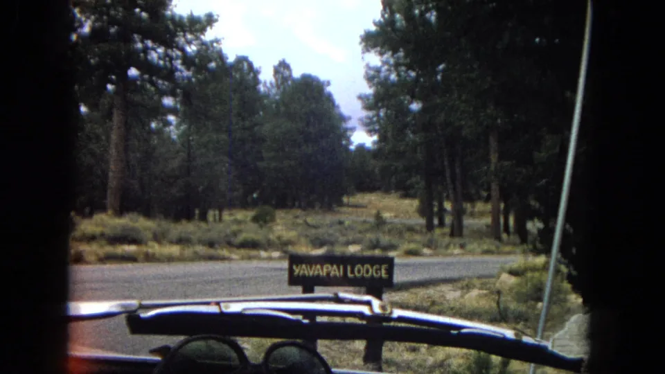 1967 home-movie frame from a road approach near Yavapai Lodge, with the lodge sign, pine trees, roadside brush, pavement, and part of a vehicle visible in the foreground.