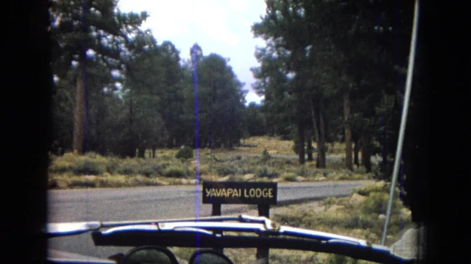 1967 Arizona 8mm frame showing a wooden Yavapai Lodge roadside sign among pine trees, filmed from a car at the lodge entrance.