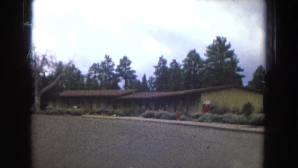 1967 Arizona 8mm frame showing a low Yavapai Lodge building set back from a paved drive with trees and shrubs around it.