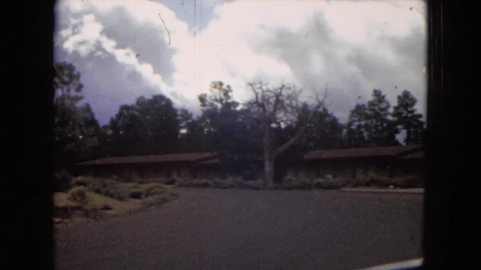 1967 Arizona 8mm frame showing a wider view of the Yavapai Lodge exterior across a paved drive, with pine trees and cloud cover behind it.