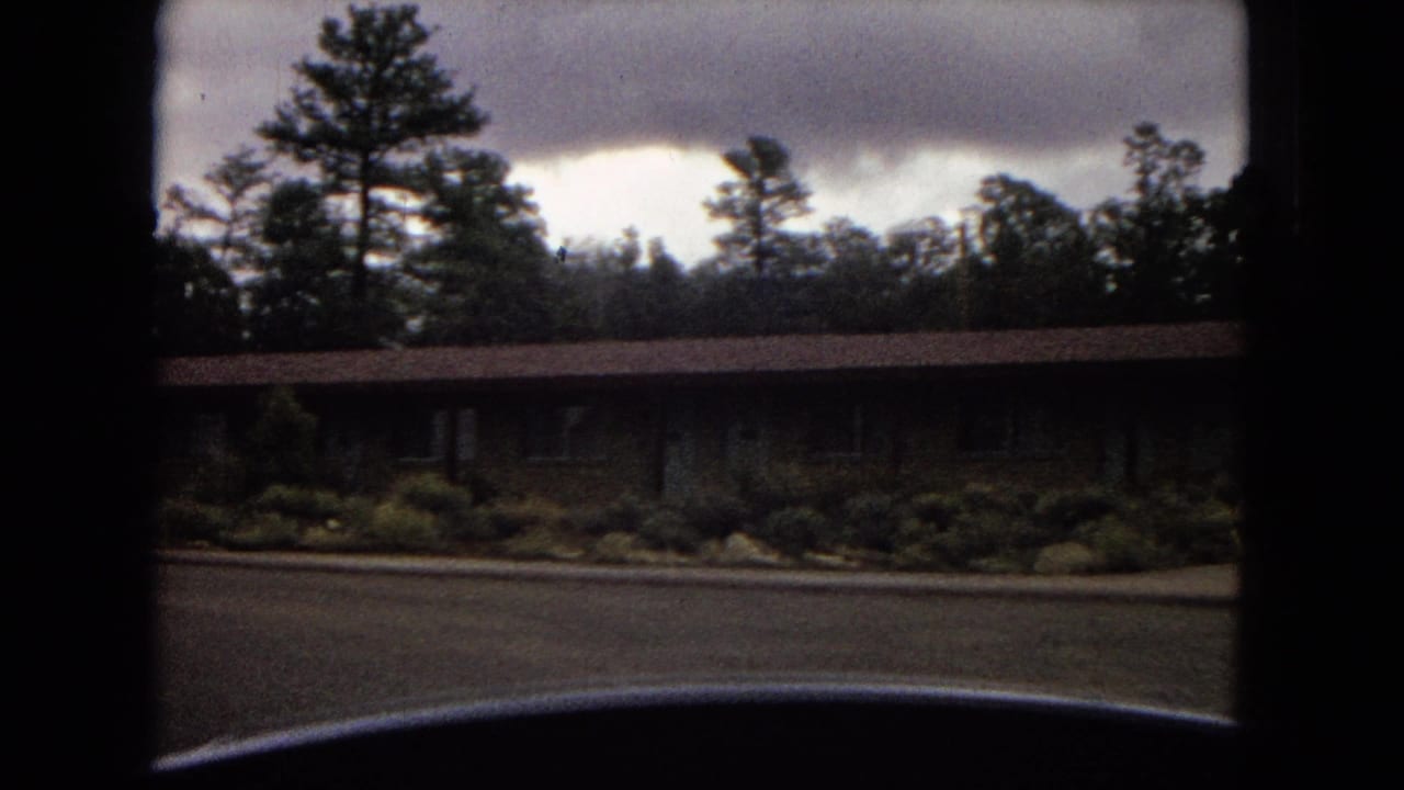 1967 Arizona home-movie frame showing a long low building with shrubs, pine trees, cloudy sky, and a dark vehicle edge at the bottom.