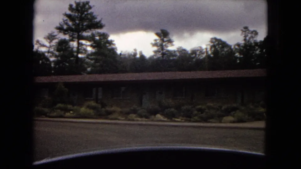 1967 Arizona home-movie frame showing a long low building with shrubs, pine trees, cloudy sky, and a dark vehicle edge at the bottom.