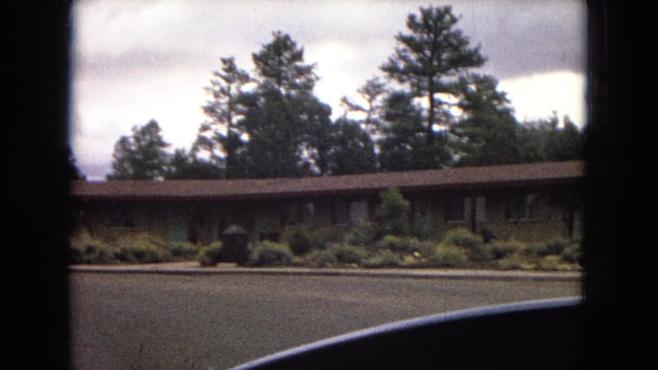 1967 Arizona travel footage frame with a low building, planted shrubs, tall pines, overcast sky, and the curved edge of a vehicle in the foreground.