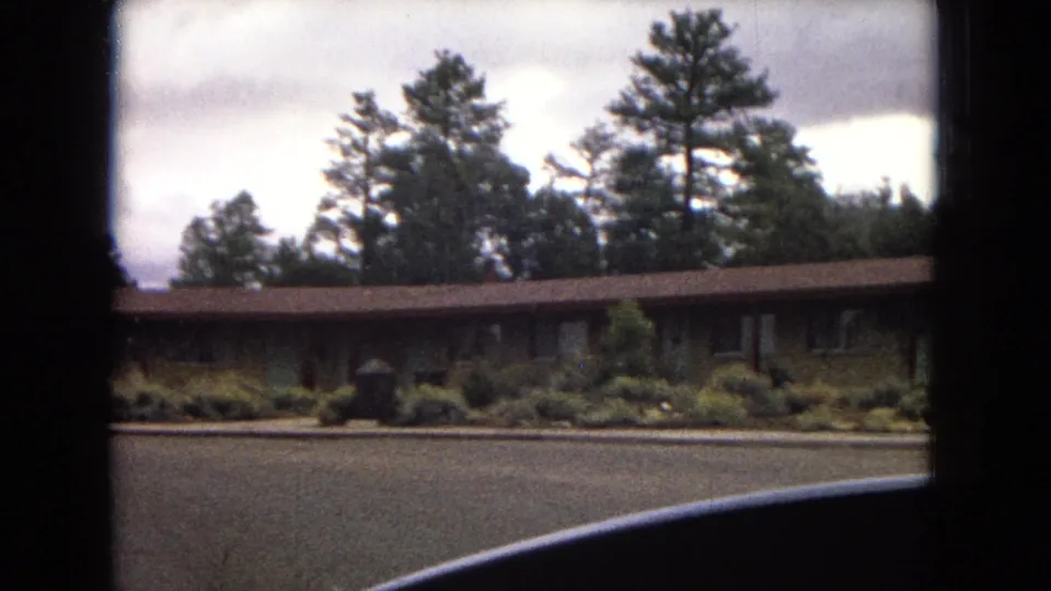 1967 Arizona travel footage frame with a low building, planted shrubs, tall pines, overcast sky, and the curved edge of a vehicle in the foreground.