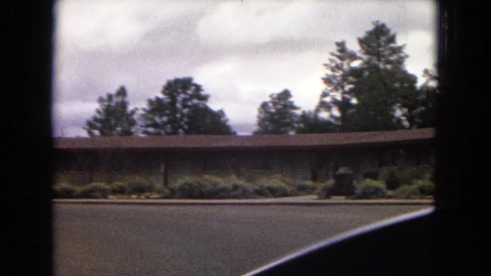 1967 Arizona home-movie frame of a long single-story roadside building beneath pine trees, with shrubs along the frontage and a dark car edge crossing the lower frame.