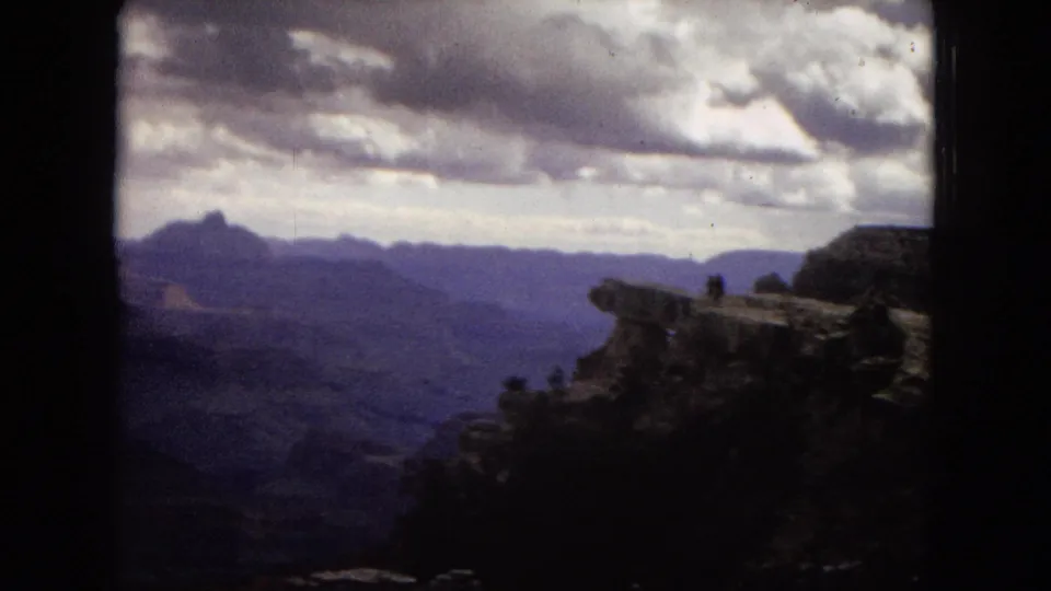 1967 Arizona frame showing a dramatic Grand Canyon overlook, layered distant cliffs, heavy clouds, and one small person standing near the cliff edge.