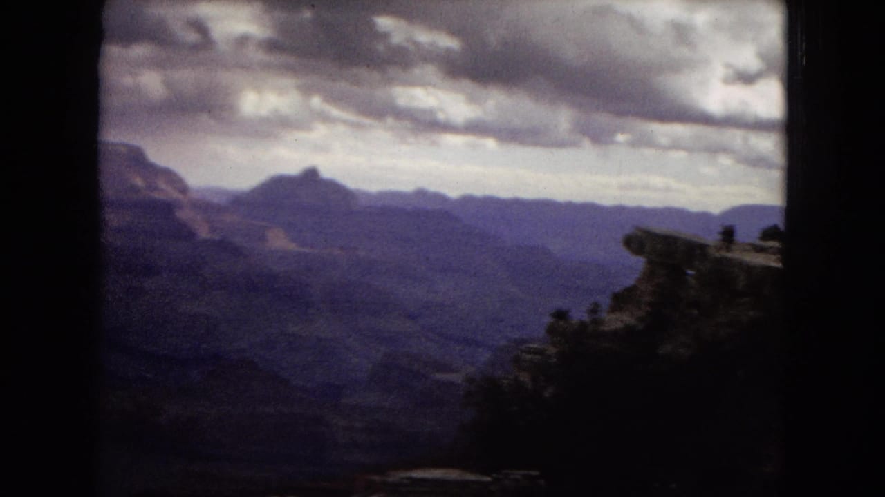 1967 8mm home-movie view of the Grand Canyon from a high overlook, with shadowed foreground rock, distant ridgelines, and heavy clouds over the canyon.