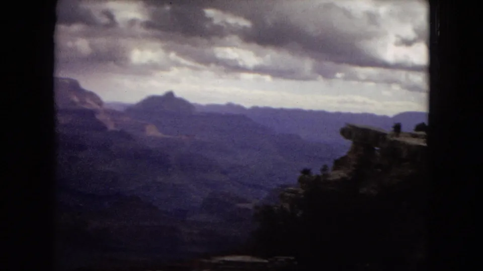 1967 8mm home-movie view of the Grand Canyon from a high overlook, with shadowed foreground rock, distant ridgelines, and heavy clouds over the canyon.