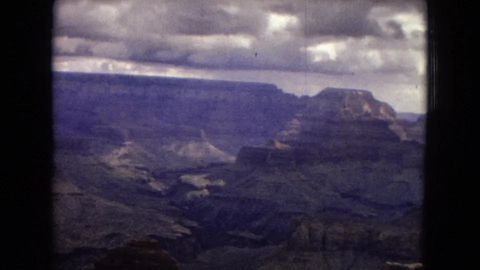 Wide 1967 Grand Canyon panorama on 8mm film, showing a broad mesa, layered canyon formations, deep ravines, and a dramatic overcast sky.