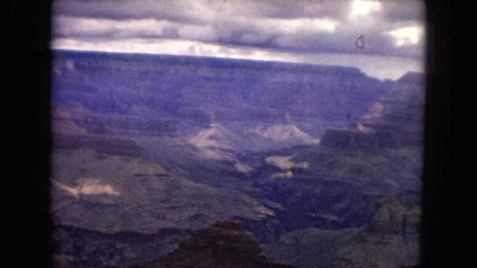 1967 8mm view across the Grand Canyon basin, with layered rock shelves, a distant canyon wall, muted blue-purple tones, and cloud shadows across the formations.