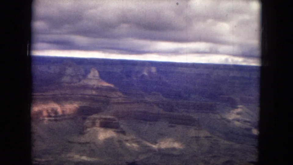 1967 8mm Grand Canyon landscape with mesa-like formations, shadowed ravines, a broad cloud band, and soft faded color across the frame.
