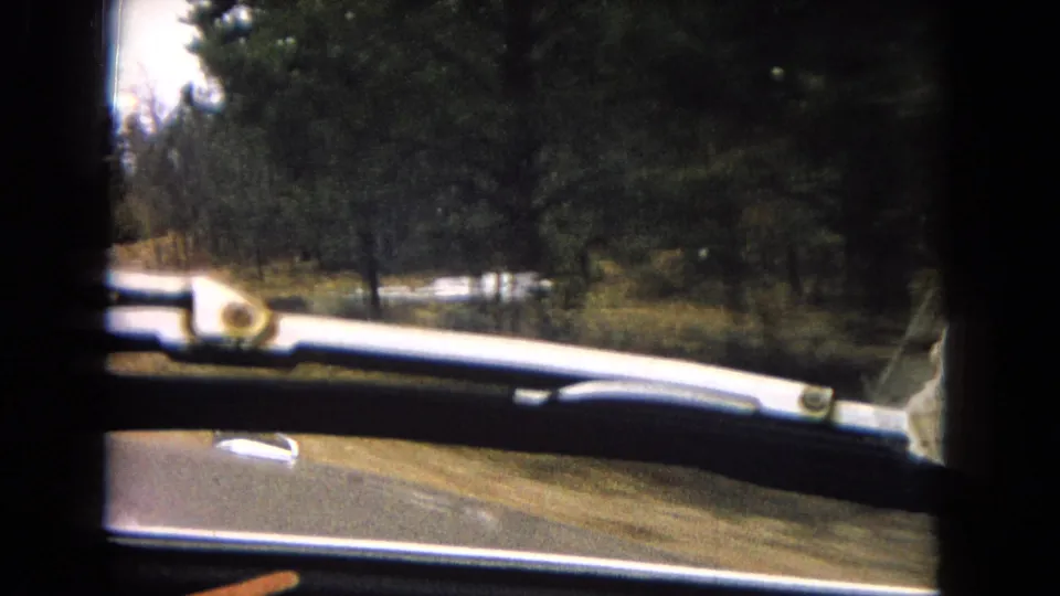 Interior vehicle viewpoint from 1967 8mm footage, with the car hood and windshield wiper visible as the road continues through pine forest.