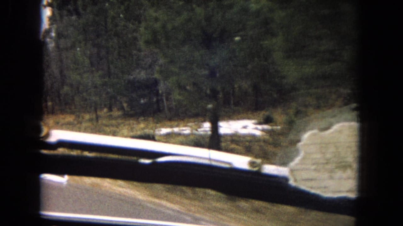 1967 Arizona home-movie frame from inside a moving car, showing a roadside curve, windshield wiper, dashboard edge, and wooded arid terrain.