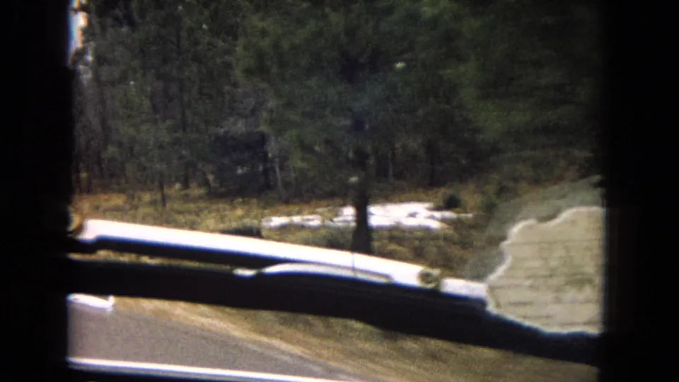 1967 Arizona home-movie frame from inside a moving car, showing a roadside curve, windshield wiper, dashboard edge, and wooded arid terrain.