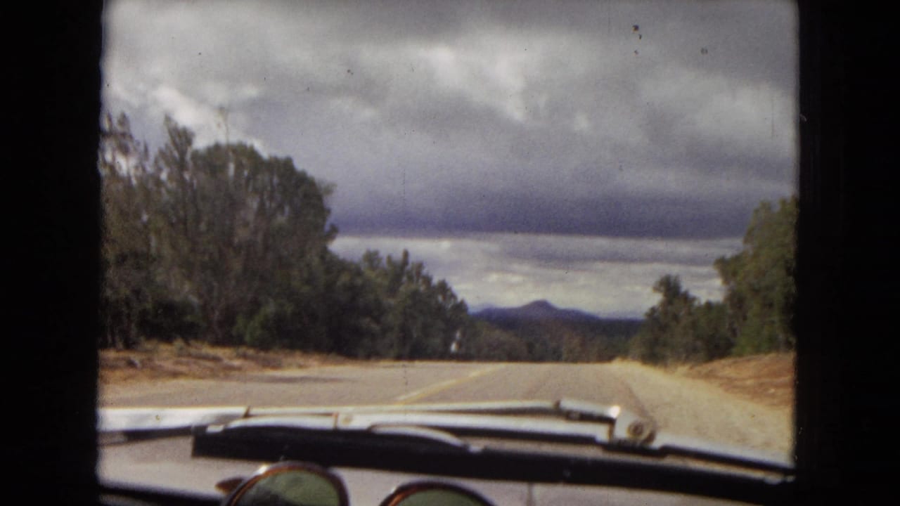 1967 Arizona driving view through the windshield, with dashboard details, sunglasses, roadside trees, distant mountains, and a dark storm cloud bank.