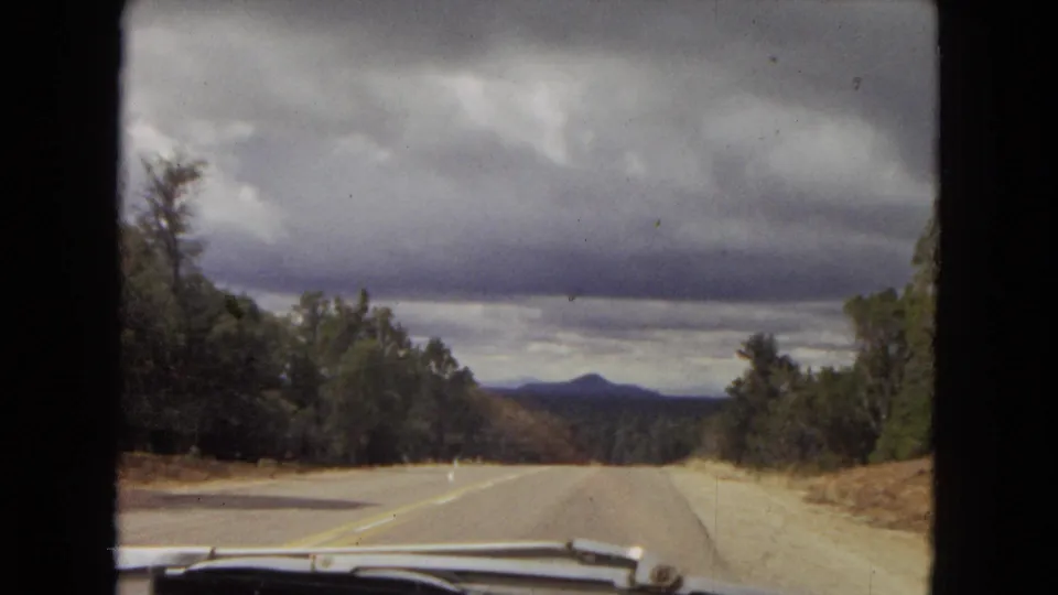 Windshield perspective from a 1967 Arizona road trip, showing the highway descending toward distant hills under layered storm clouds.