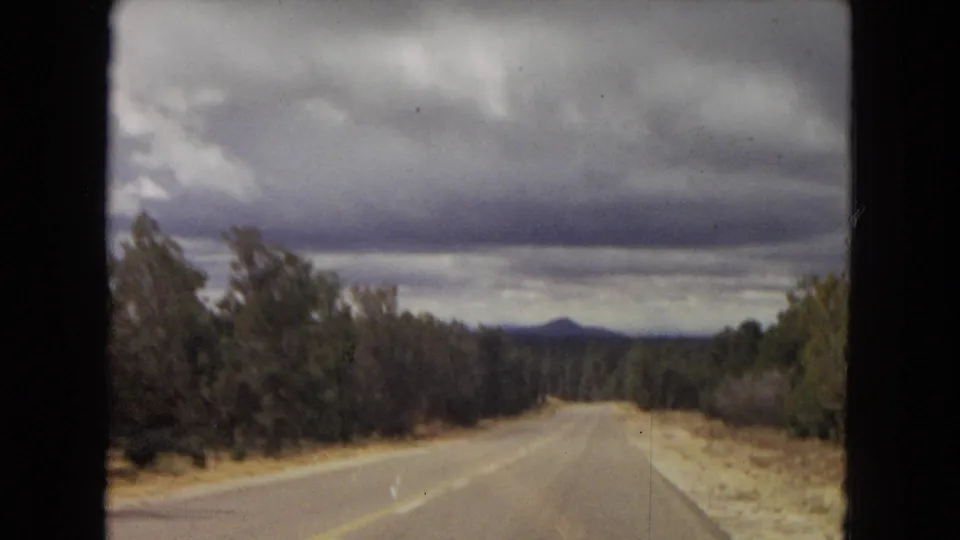 Final 1967 Arizona windshield view of a rural road bordered by trees and pale roadside soil, with distant mountains beneath heavy clouds.