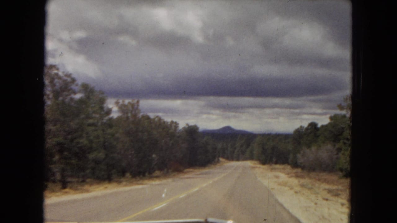1967 Arizona home-movie road view from a moving vehicle, showing a two-lane road bordered by trees and scrub beneath heavy clouds.