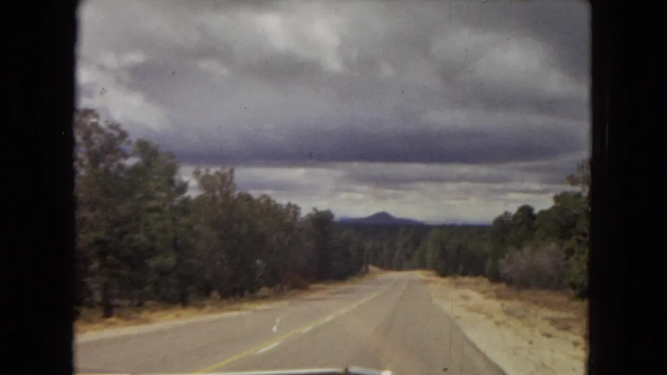 1967 Arizona home-movie road view from a moving vehicle, showing a two-lane road bordered by trees and scrub beneath heavy clouds.
